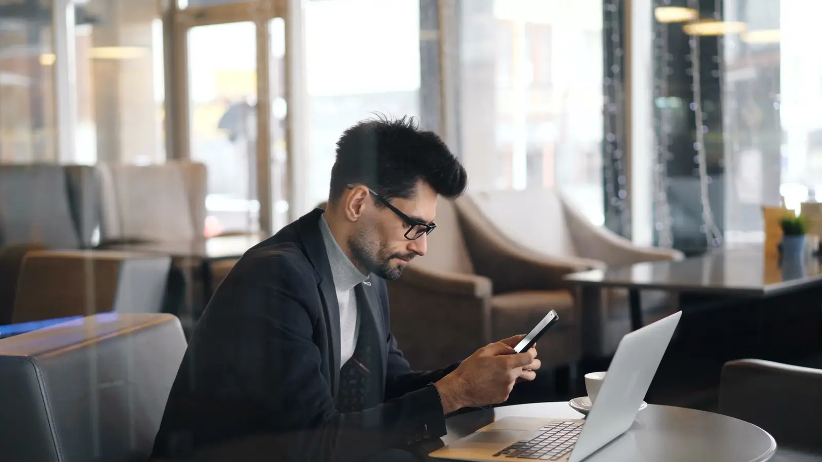 Personne dans un café, téléphone en main, ordinateur ouvert sur la table.