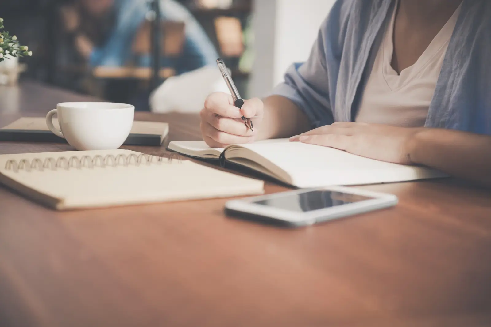 Personne qui écrit dans un cahier, un téléphone posé sur la table.